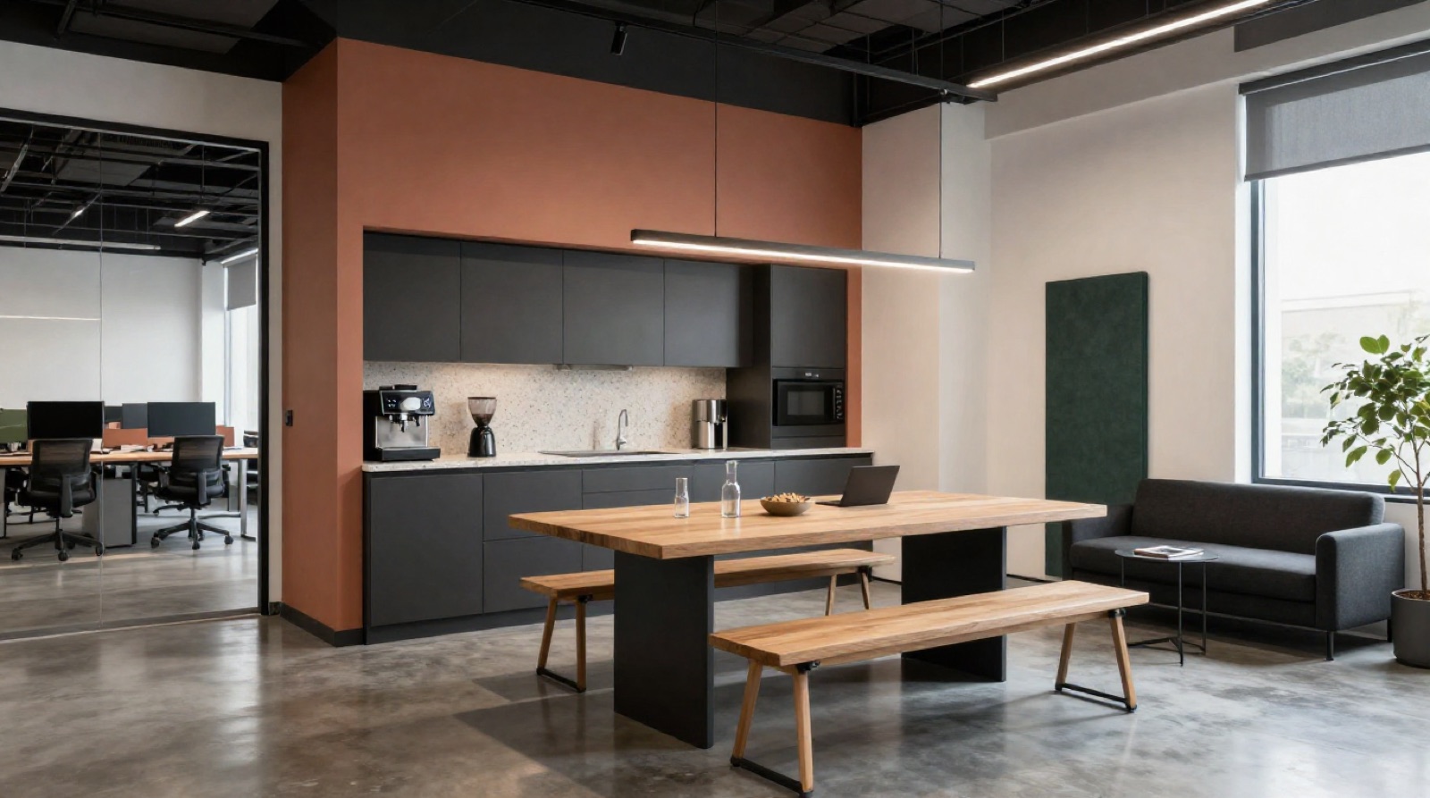 Comedor y office de una oficina tecnológica en Madrid con pared ancla en terracota suave detrás de la kitchenette en charcoal mate con encimera terrazo, cafetera profesional, mesa comunal de roble con bancos, lámpara lineal de techo, panel acústico en fieltro verde y butaca modular a un lado, techo expuesto pintado de negro y partición de cristal al otro lado que deja ver la zona de trabajo, con luz natural diurna lateral
