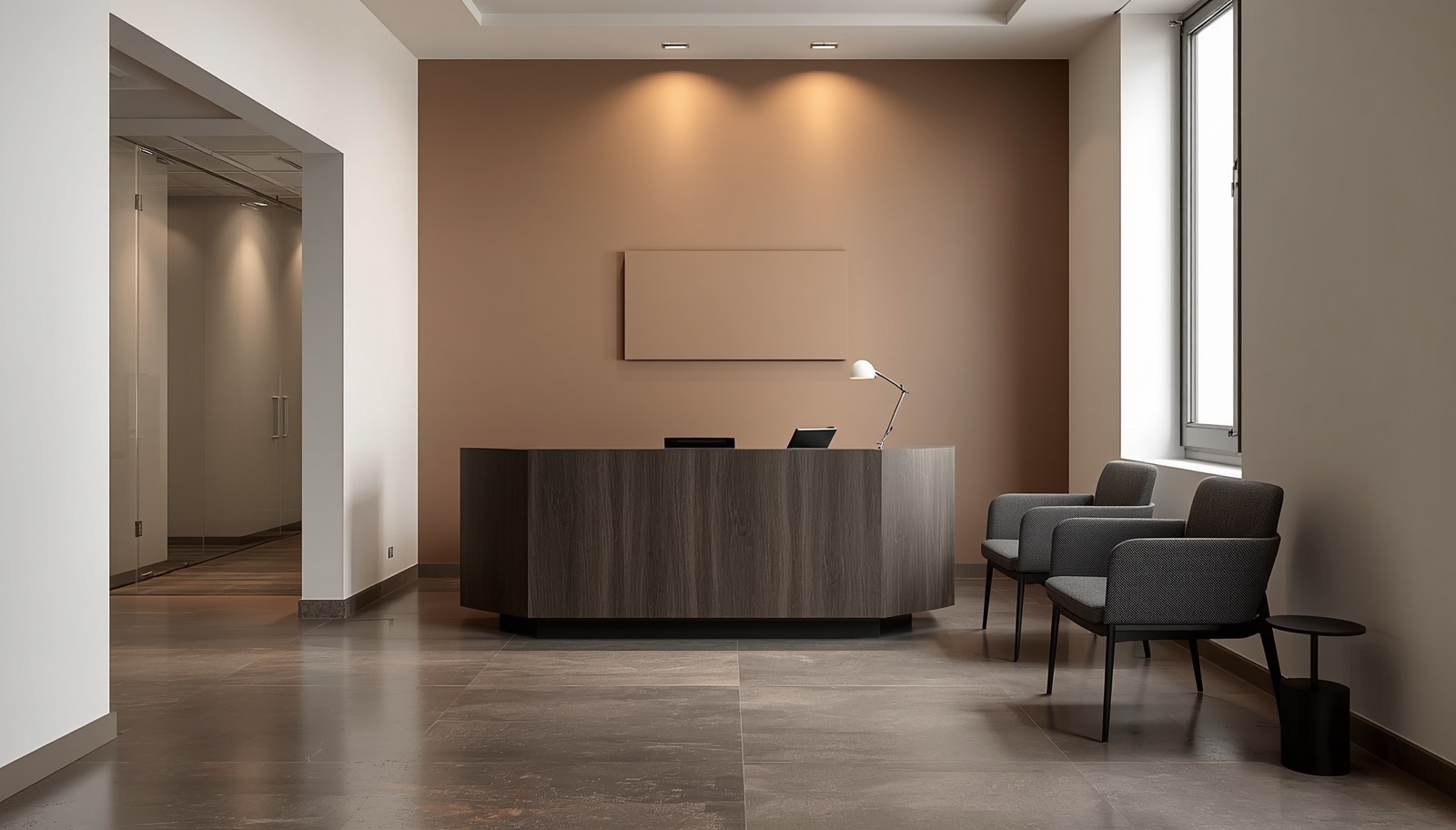 Recepción corporativa de una oficina en Madrid con mostrador bajo de madera oscura mate, pared ancla pintada en terracota apagada detrás y paredes laterales en blanco roto cálido; a un lado, partición de cristal con franja de privacidad que deja ver un pasillo con puertas; al otro, zona de espera con dos butacas grises y mesa auxiliar negra, iluminada con luz natural diurna